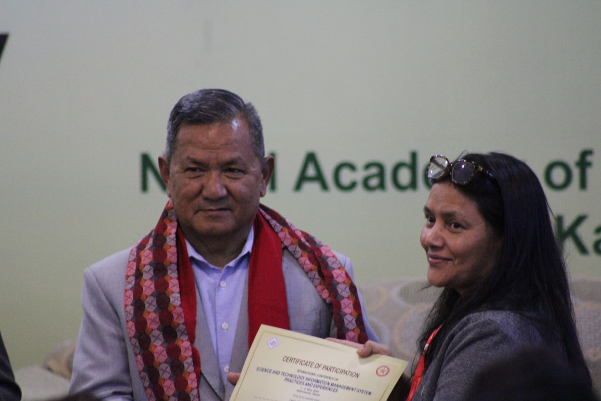 A man in a grey suit and red patterned scarf hands a certificate of participation to a woman in a dark top, with a green logo visible in the background.