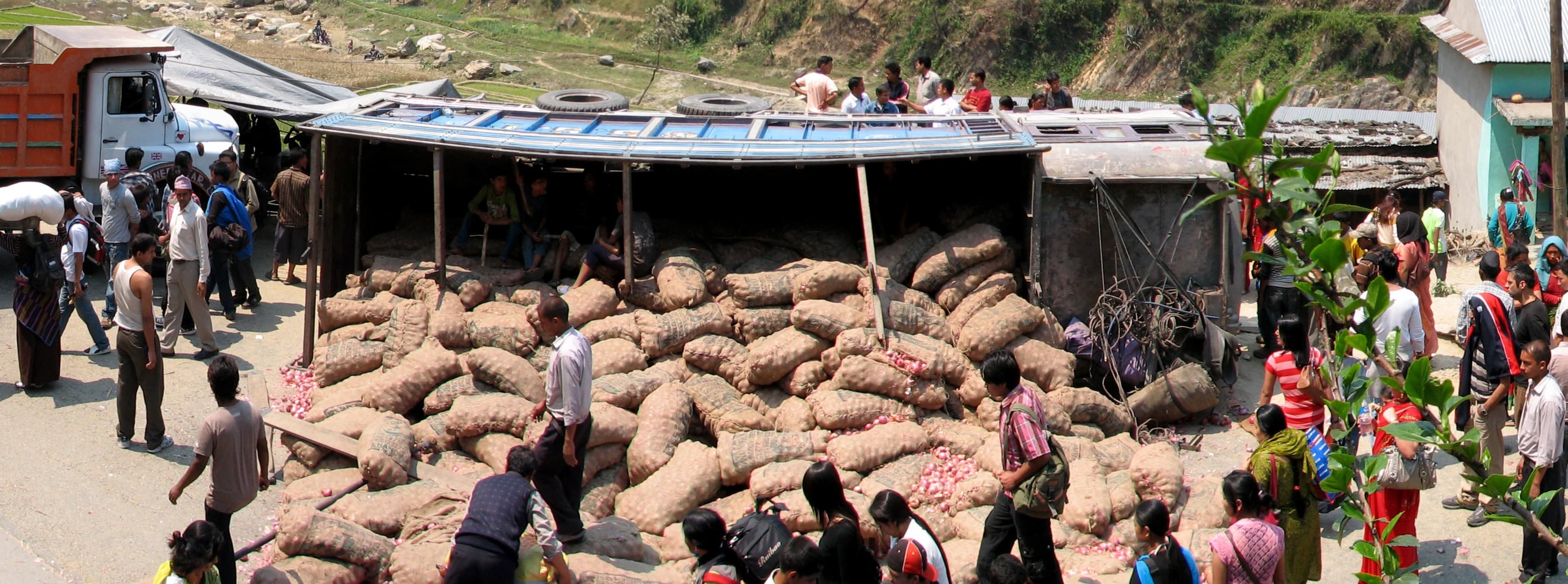 A wide view of a chaotic roadside accident scene with an overturned truck, scattered goods, an orange truck, and numerous onlookers gathered on a dusty road in a rural, hilly area.