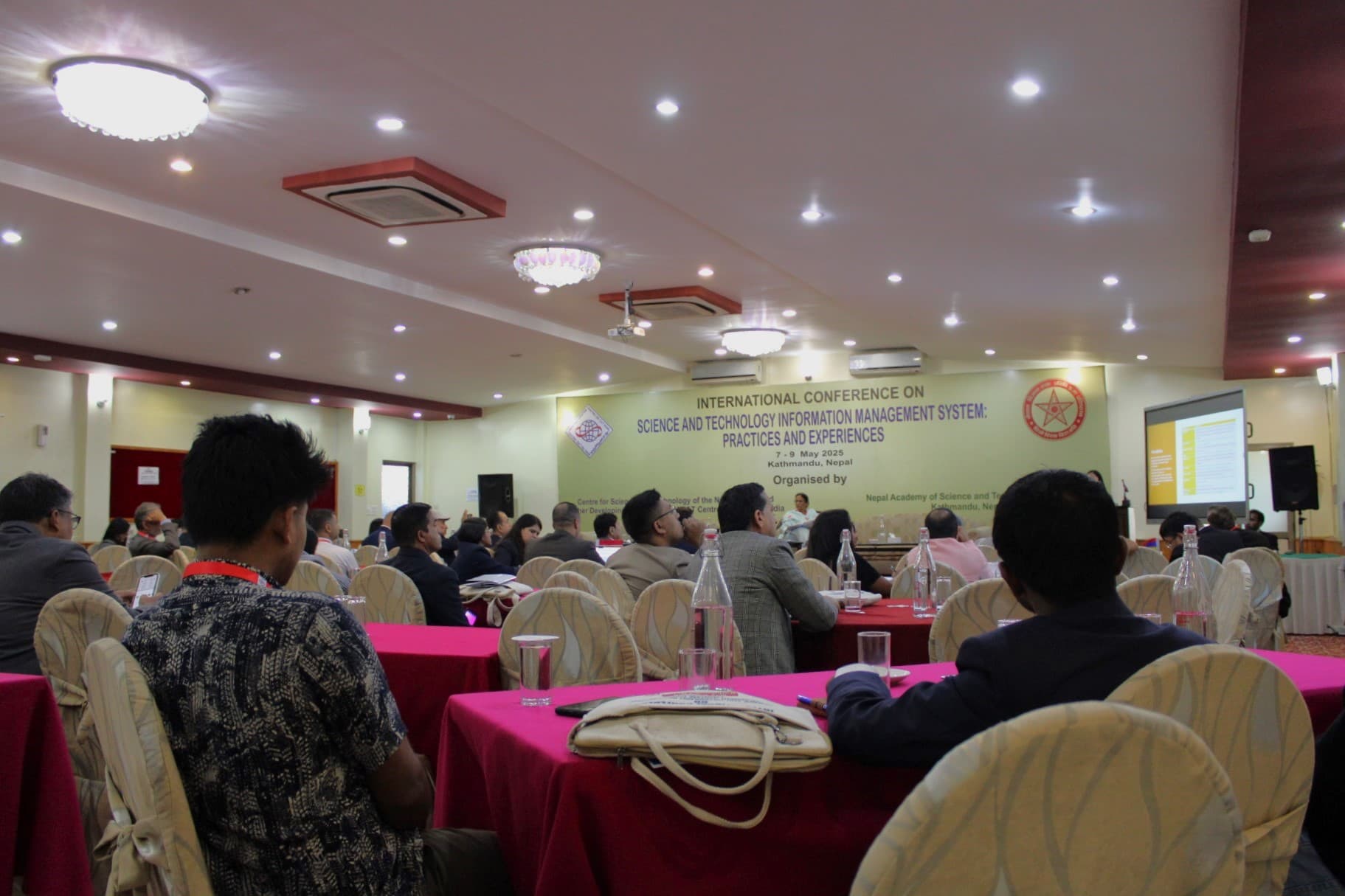 A wide shot of a conference room with many attendees seated at tables with pink tablecloths, facing a stage with a banner that reads "INTERNATIONAL CONFERENCE ON SCIENCE AND TECHNOLOGY INFORMATION MANAGEMENT SYSTEM: PRACTICES AND EXPERIENCES".